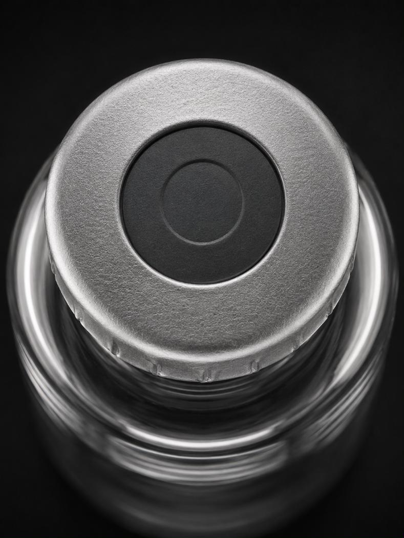 Overhead close-up of a sealed vial against a dark ground: a brushed aluminium crimp seal with a matte dark grey flip-off disc at its centre, the crimp folds visible around the rim.